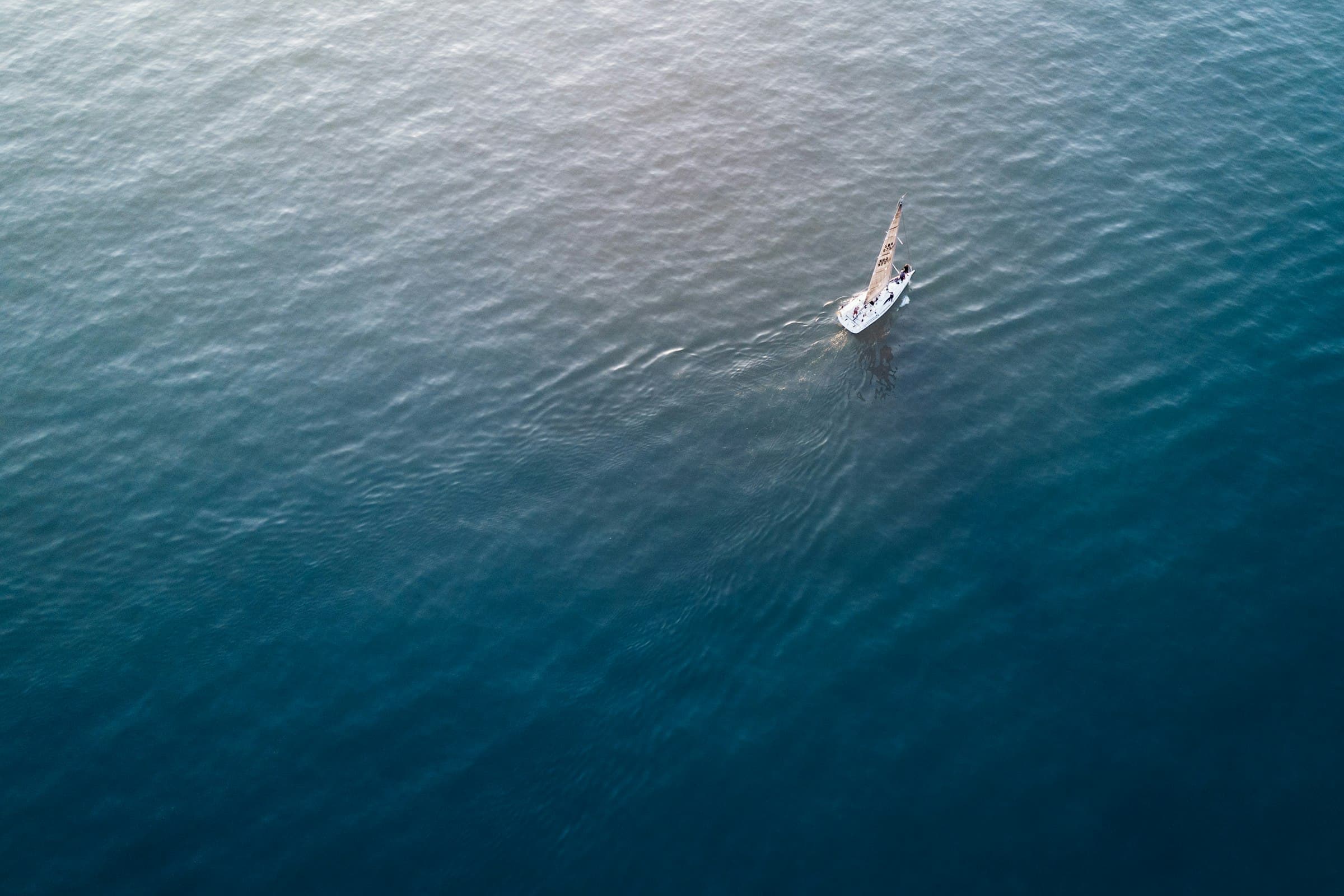 Aerial view of a white sailboat crossing calm open water, its wake trailing behind